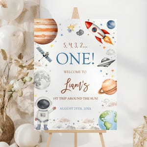 May include: A wooden easel displays a colourful space-themed sign. The sign features planets, a rocket, and an astronaut illustration. Text includes "ONE!" and "Liam's 1st Trip Around the Sun!" with a date. Balloons and decorative elements surround the sign.