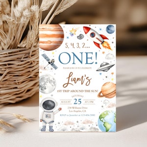 May include: A blue and white space-themed first birthday party invitation with a rocket, planets, stars, and an astronaut. The text reads "5, 4, 3, 2... ONE! Please join us to celebrate Liam's 1st trip around the sun! Saturday, August 25 at 2 PM, 234 Williams Drive, Los Angeles, CA. RSVP to Jennifer at 123-456-7890."