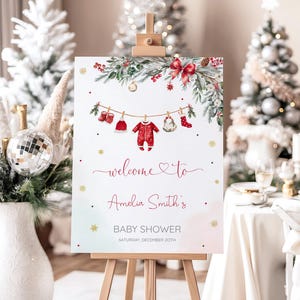 May include: A Christmas-themed baby shower sign on an easel. The sign displays a watercolour design with greenery, red accents, and baby clothes hanging on a line. Text reads "welcome to Amelia Smith's BABY SHOWER SATURDAY, DECEMBER 20TH."