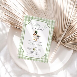 May include: Baby shower invitation with a green gingham border. The invitation features a watercolor illustration of a duck and the text "A Baby Boy Is On His Way!" with event details. The invitation is on a white plate with a dried palm leaf.