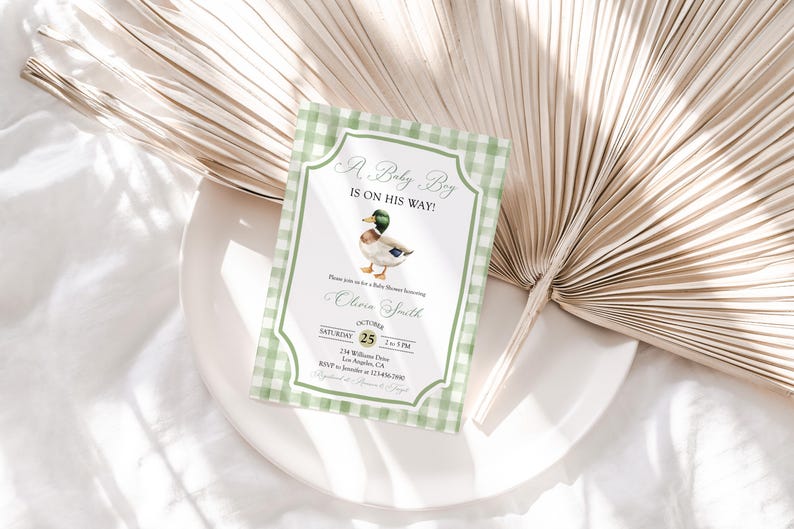 May include: Baby shower invitation with a green gingham border. The invitation features a watercolor illustration of a duck and the text "A Baby Boy Is On His Way!" with event details. The invitation is on a white plate with a dried palm leaf.