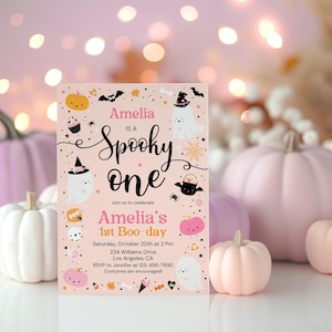 以下が含まれることがあります： 「Amelia is a Spooky One」と「Amelia's 1st Boo-day」のテキストが入った、ピンク色のハロウィーンをテーマにした招待状。カボチャ、幽霊、コウモリ、その他のハロウィーンの要素のイラストが特徴です。背景にはピンク色のカボチャがあります。