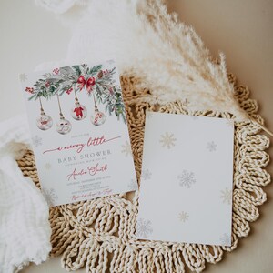 May include: A Christmas-themed baby shower invitation. The design includes watercolour ornaments, foliage, and a red bow. The text reads "a merry little baby shower". A blank card with snowflake patterns is also visible, all on a neutral background.