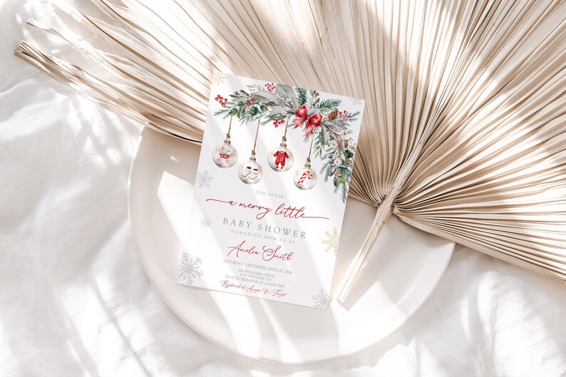 May include: A Christmas-themed baby shower invitation. The card features festive illustrations of baubles, greenery, and snowflakes. The text reads "a merry little baby shower". The invitation is on a white plate.