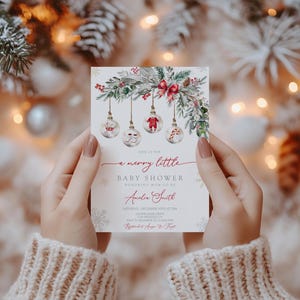 May include: A Christmas-themed baby shower invitation. The card, held by a person, features a festive design with hanging baubles, greenery, and the text "a merry little baby shower". The invitation is decorated with snowflakes.