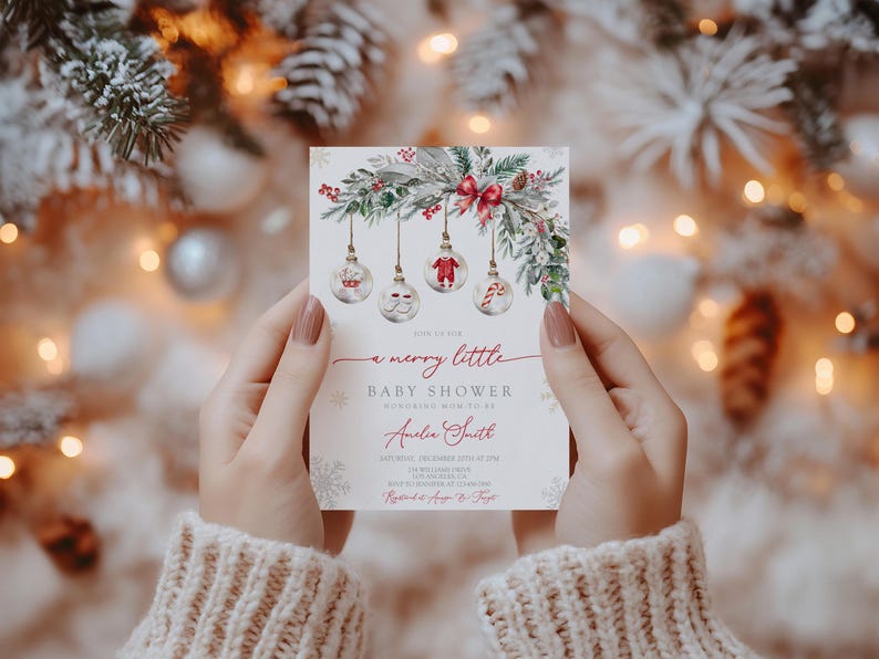 May include: A Christmas-themed baby shower invitation. The card, held by a person, features a festive design with hanging baubles, greenery, and the text "a merry little baby shower". The invitation is decorated with snowflakes.