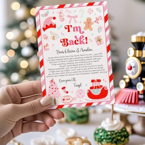 May include: A Christmas-themed card held by a hand, featuring a pink and red striped border. The card has the text "I'm Back!" and a letter to Olivia & Amelia from their Elf, Tinsel, with festive illustrations.