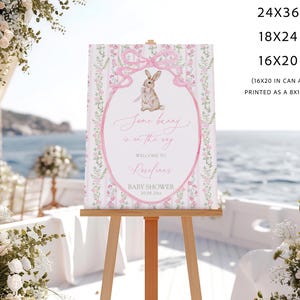 May include: A baby shower sign on an easel. The sign features a pink oval with a bunny illustration and the text "Some bunny is on the way." The sign is surrounded by floral and bow designs. The sign is available in 24x36, 18x24, and 16x20 inch sizes.