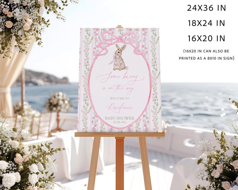 May include: A baby shower sign on an easel. The sign features a pink oval with a bunny illustration and the text "Some bunny is on the way." The sign is surrounded by floral and bow designs. The sign is available in 24x36, 18x24, and 16x20 inch sizes.