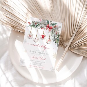 May include: A Christmas-themed baby shower invitation with the words "santa baby" in elegant script. The invitation features watercolor illustrations of ornaments, greenery, and snowflakes, with event details in a delicate font.