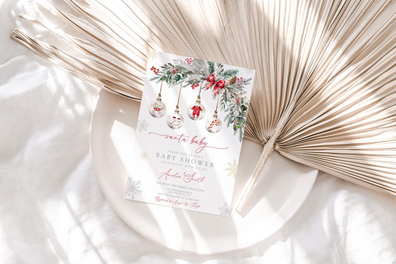 May include: A Christmas-themed baby shower invitation with the words "santa baby" in elegant script. The invitation features watercolor illustrations of ornaments, greenery, and snowflakes, with event details in a delicate font.