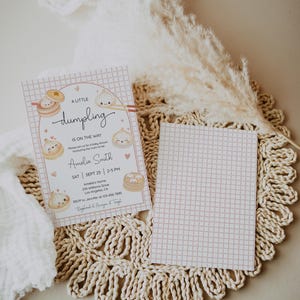 May include: A baby shower invitation with a pink grid pattern and dumpling illustrations. The text reads "A Little Dumpling is on the Way" with event details. The invitation is on a woven placemat with a decorative dried plant.