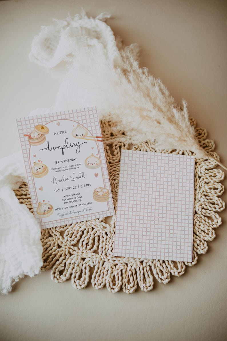 May include: A baby shower invitation with a pink grid pattern and dumpling illustrations. The text reads "A Little Dumpling is on the Way" with event details. The invitation is on a woven placemat with a decorative dried plant.