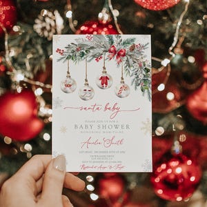 May include: A baby shower invitation with a Christmas theme. The invitation features a festive design with hanging ornaments, greenery, and a red bow. The text reads "santa baby" and includes details for the event.