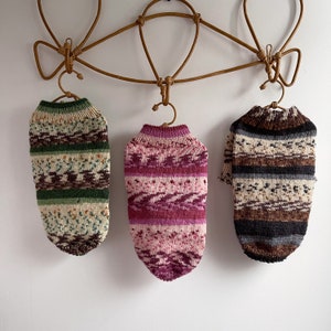 May include: Three knitted sweaters in different colors and patterns hanging on a wooden hanger. The sweaters are green and brown, pink and white, and brown and gray.