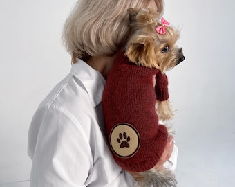 May include: A woman holding a small brown and white Yorkshire Terrier wearing a maroon sweater with a brown paw print on the left sleeve.