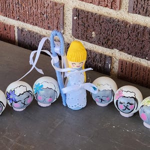 May include: A collection of hand-painted Easter egg ornaments. The eggs feature sheep faces with floral accents. A small doll with yellow hair and a blue shepherd's crook are also present. The ornaments are arranged on a dark surface against a brick background.