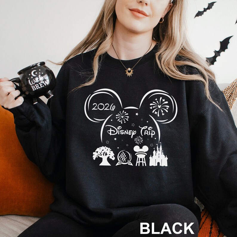 A woman wearing a black sweater with a white graphic design featuring Disney-themed elements, such as Mickey Mouse ears and fireworks, holding a black mug with the text "Disney Trip" printed on it.