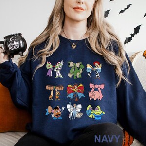 A woman wearing a navy blue sweatshirt with various cartoon characters printed on it, including characters from the Toy Story franchise. She is holding a black mug with the words "Toy Story Brew" printed on it.