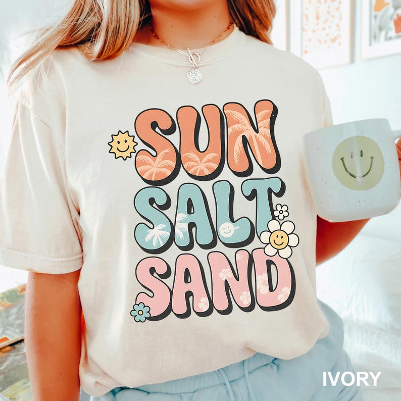 A woman wearing a white t-shirt with a colorful graphic design that says "SUN SALT SAND" and holding a white mug with a smiley face on it.