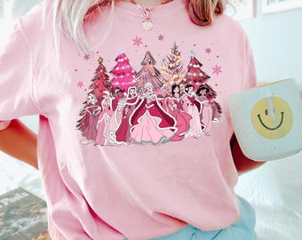Puede incluir: Camiseta rosa con un gráfico de princesas Disney de pie frente a árboles de Navidad. Las princesas visten vestidos rosas y rojos y los árboles están decorados con luces y adornos.