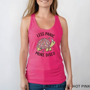 May include: A hot pink tank top featuring a graphic of a turtle with a disco ball shell wearing a party hat. The text above the turtle reads LESS PANIC and the text below reads MORE DISCO in black lettering, accompanied by small star and music note icons.