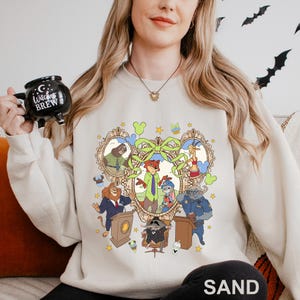 A woman wearing a white sweatshirt with a cartoon character design, holding a mug with a witch's brew logo.