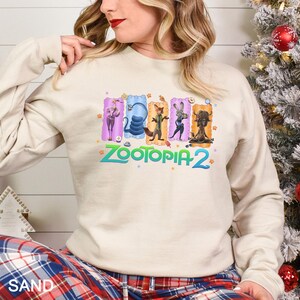 A woman wearing a white sweatshirt with a colorful graphic of five cartoon animal characters from the movie "Zootopia 2" on it.