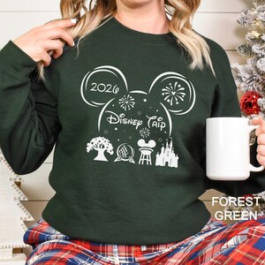 A person wearing a green sweater with a white design featuring Disney-themed graphics and text, holding a white mug.