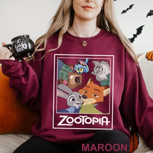 May include: Maroon sweatshirt featuring a Zootopia graphic. The graphic includes characters from the animated movie, such as Judy Hopps and Nick Wilde. The word "Zootopia" is printed in white. The word "MAROON" is printed at the bottom.