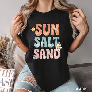A woman wearing a black t-shirt with the text "SUN SALT SAND" printed on it.