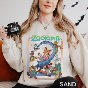 A woman wearing a white sweatshirt with a cartoon character design and holding a mug with a witch's brew logo.