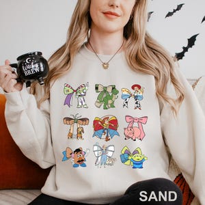 A woman wearing a white sweatshirt with a graphic of various cartoon characters printed on it, holding a mug with a witch's brew design.