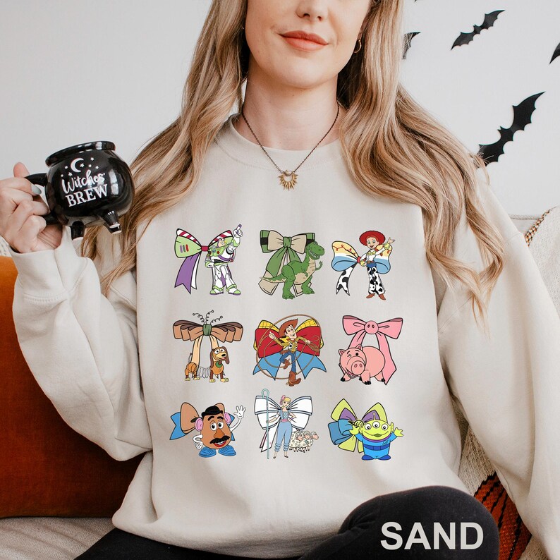 A woman wearing a white sweatshirt with a graphic of various cartoon characters printed on it, holding a mug with a witch's brew design.