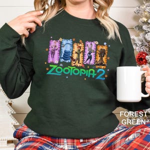 A woman wearing a green sweatshirt with a colorful graphic of cartoon animals and the text "Zootopia 2" printed on it, holding a white mug.