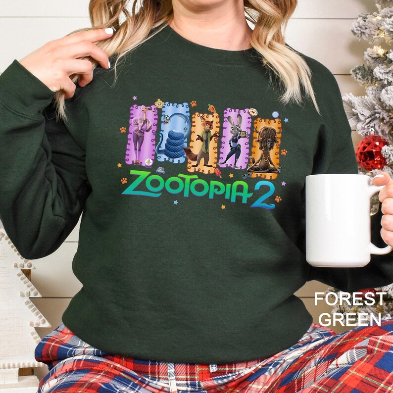 A woman wearing a green sweatshirt with a colorful graphic of cartoon animals and the text "Zootopia 2" printed on it, holding a white mug.