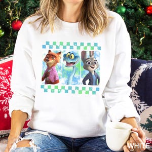 A woman wearing a white sweatshirt with a graphic of three cartoon animal characters - a fox, a snake, and a rabbit - on it. She is sitting on a couch and holding a white mug.