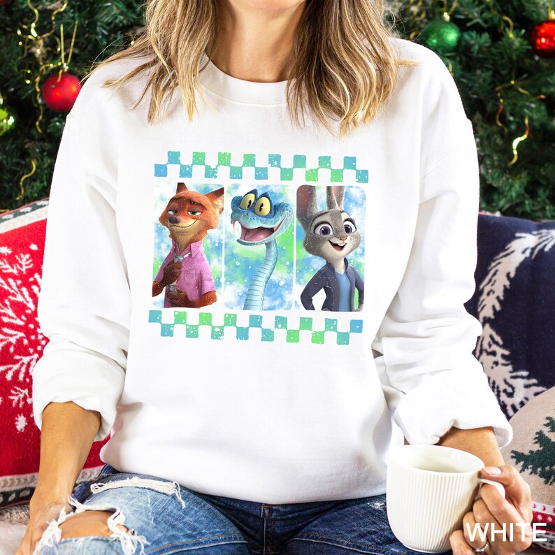 A woman wearing a white sweatshirt with a graphic of three cartoon animal characters - a fox, a snake, and a rabbit - on it. She is sitting on a couch and holding a white mug.