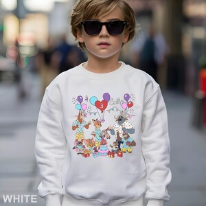 A young boy wearing a white sweatshirt with a colorful cartoon character design and a birthday theme.