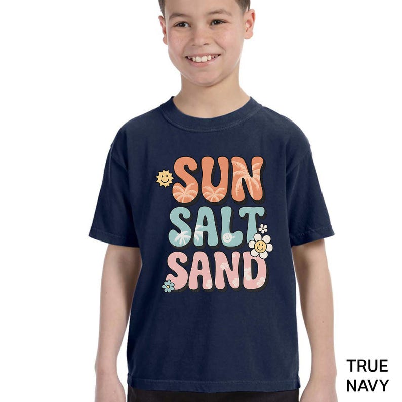 A young boy wearing a navy blue t-shirt with the text "SUN SALT SAND" printed on it.