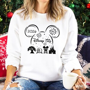 A woman wearing a white sweater with a graphic design featuring a Mickey Mouse-like symbol and various Disney-related icons, including a castle, tree, and fireworks. She is sitting on a couch with a mug in her hand.