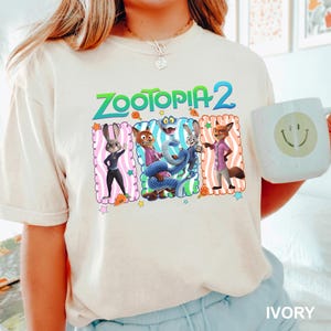 A person wearing a white t-shirt with a graphic of the Zootopia 2 characters and holding a coffee mug with a smiley face.