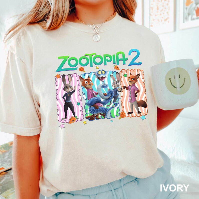 A person wearing a white t-shirt with a graphic of the Zootopia 2 characters and holding a coffee mug with a smiley face.