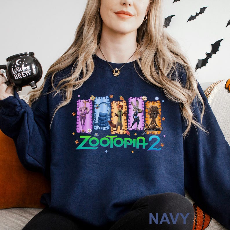 A woman wearing a navy blue sweatshirt with a colorful graphic design featuring characters from the animated film "Zootopia 2". She is holding a black mug with a Halloween-themed design.