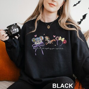 A woman wearing a black sweater with a graphic design of cartoon characters and text, holding a black mug with the text "You can do anything you wish to be" printed on it.