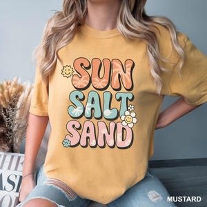 A woman wearing a yellow t-shirt with the text "SUN SALT SAND" printed on it.