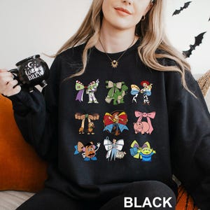 A woman wearing a black sweatshirt with a graphic of various cartoon characters printed on it, holding a black mug with the text "Witch Brew" printed on it.