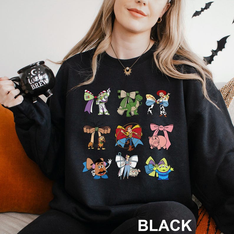 A woman wearing a black sweatshirt with a graphic of various cartoon characters printed on it, holding a black mug with the text "Witch Brew" printed on it.