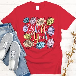 May include: A red t-shirt with a colorful seashell design and the words "Shell Yeah". The shirt is paired with blue jeans and gray sneakers, creating a casual, beach-themed ensemble.