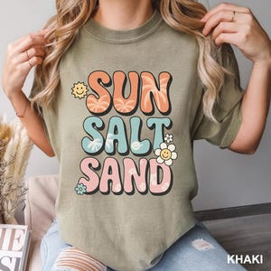 A woman wearing a green t-shirt with a colorful graphic design that says "SUN SALT SAND" and includes various beach-themed elements such as a sun, palm trees, and flowers.